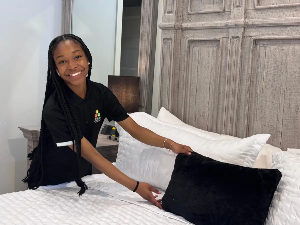 A Dunlap cleaner in a branded polo setting pillows on a freshly made bed during a turnover