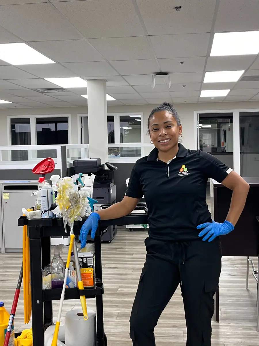 A Dunlap cleaner in gloves beside a stocked cleaning cart in an open-plan office